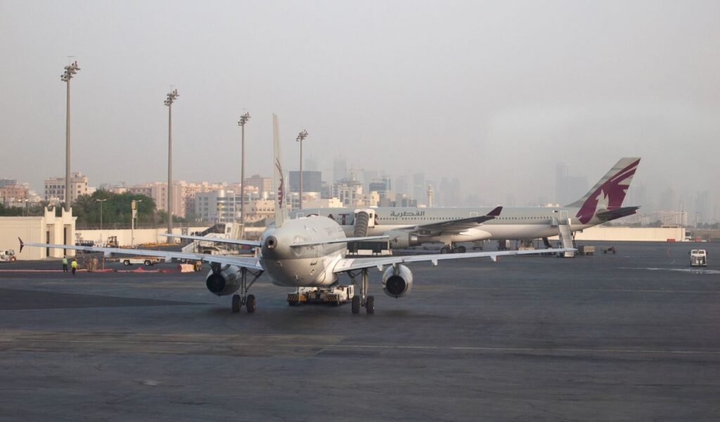 Qatar Airways aircraft parked on the tarmac with ground crew and hazy Doha city skyline visible in the background.© Vyacheslav Argenberg